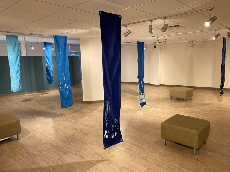 A view of Liz Miller's exhibit in the Regier Art Gallery: long vertical strips of fabric in various shades of blue with representations of the sounds in white
