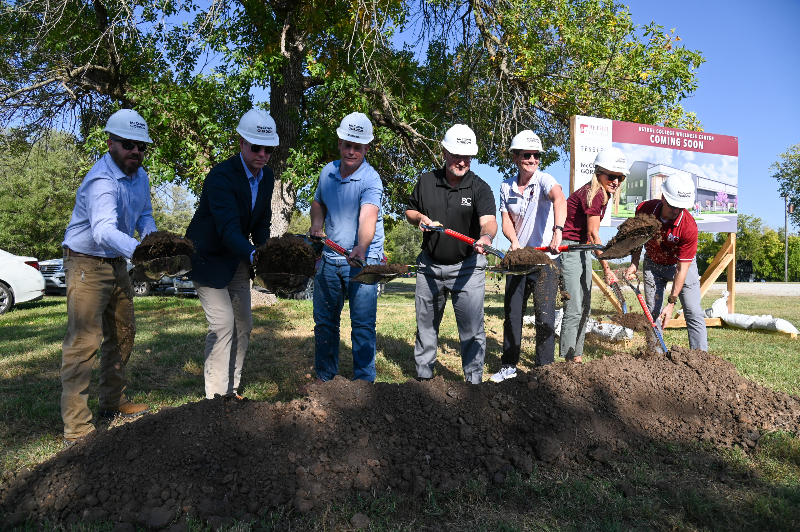 Seven people in white hardhats with shovels break ground for the new Wellness Center, pictured on a sign behind them.