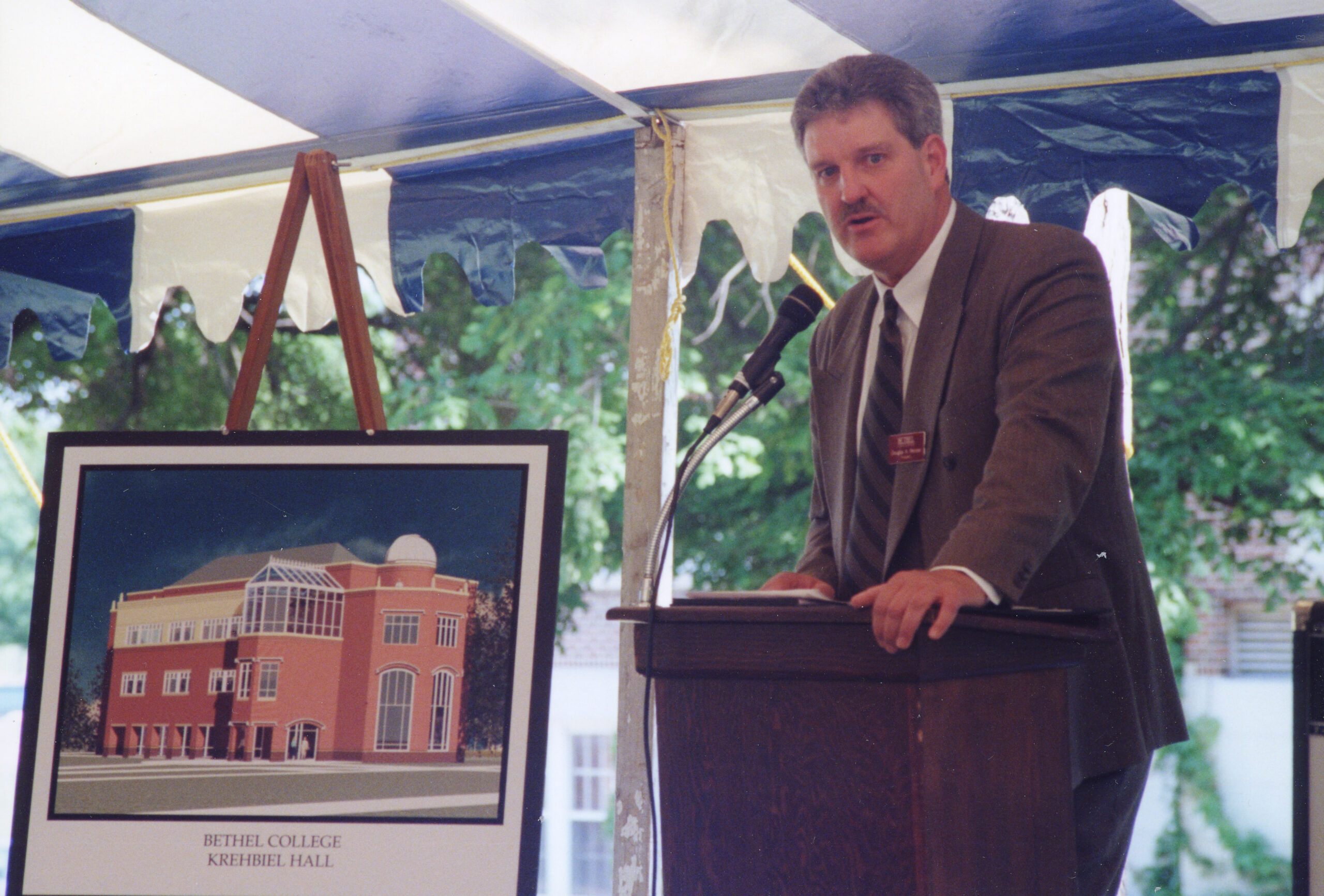 A 2000 photo of Doug Penner at the groundbreaking for Krehbiel Science Center; standing behind a podium, with an artist's rendering of the proposed building on an easel beside him.