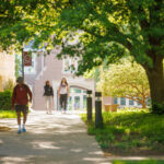 View down a partially shaded sidewalk with trees on one side, looking toward Krehbiel Science Center, with three people walking on the sidewalk