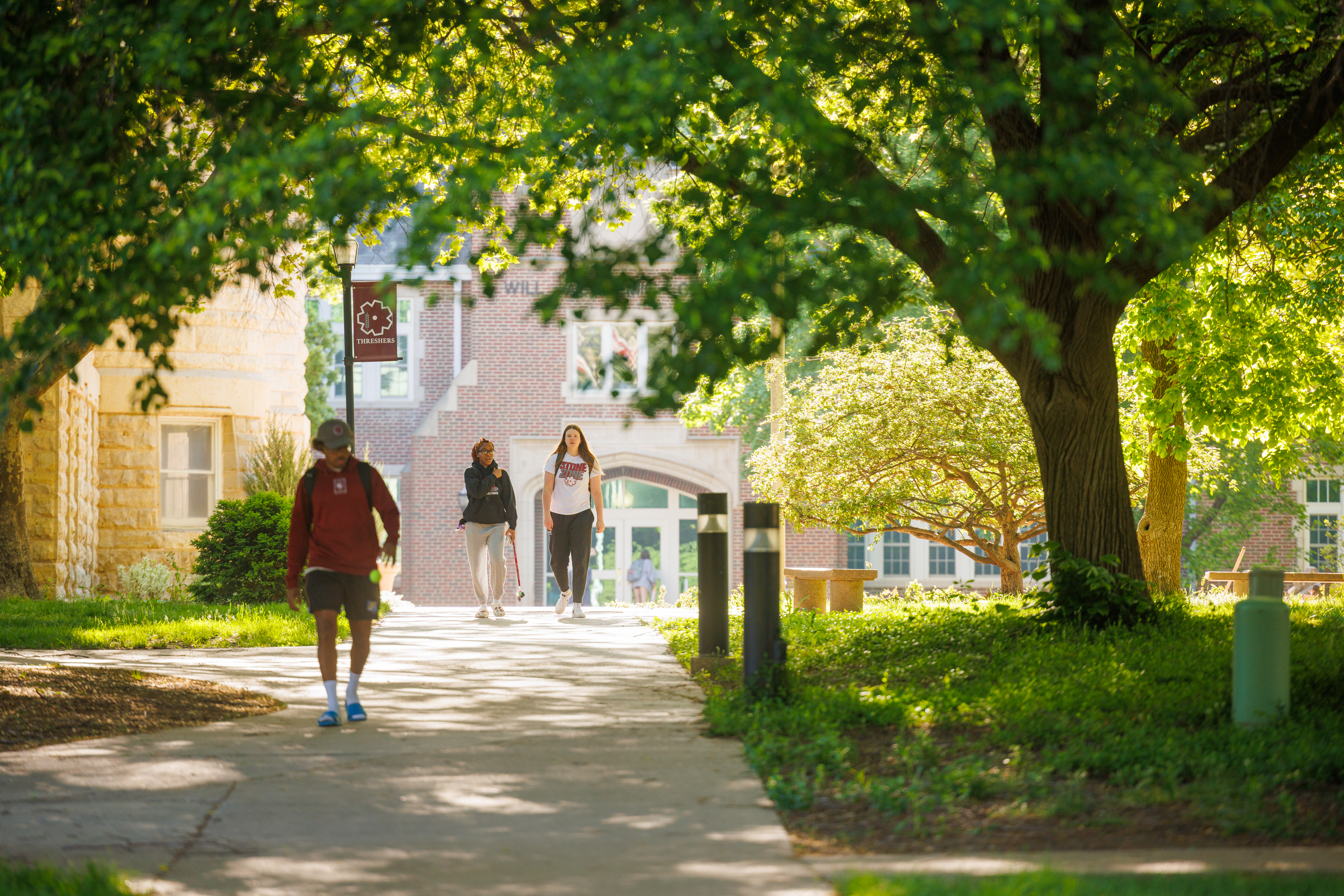 View down a partially shaded sidewalk with trees on one side, looking toward Krehbiel Science Center, with three people walking on the sidewalk
