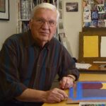 Bob Regier in his home studio: An older white man with white hair, wearing a dark-colored shirt, with in-progress artwork and supplies around him.