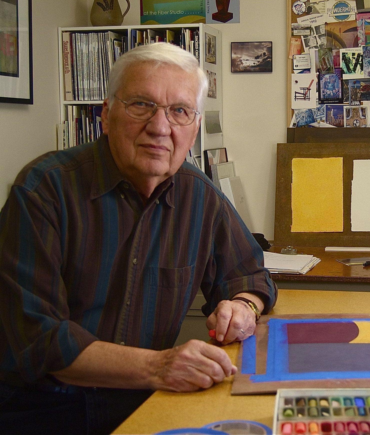 Bob Regier in his home studio: An older white man with white hair, wearing a dark-colored shirt, with in-progress artwork and supplies around him.