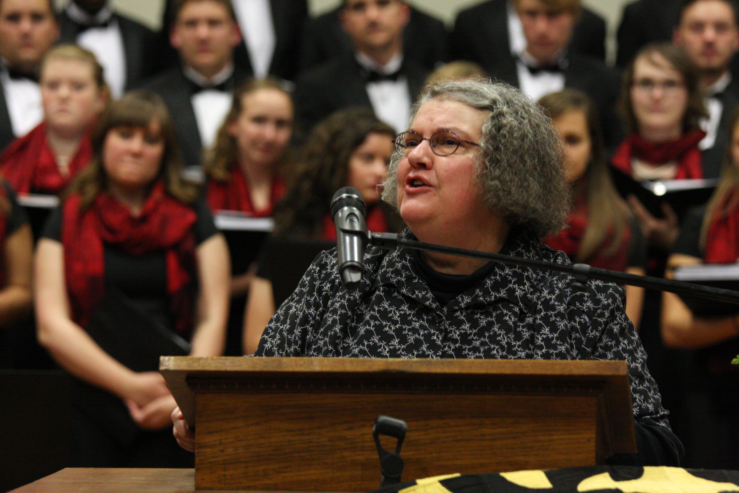 Patty Shelly speaking in Memorial Hall in 2015, standing at the large wooden podium with choir members behind her