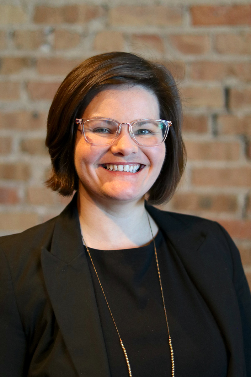 Headshot of woman with short brown hair and glasses, wearing a dark-colored jacket and shirt, standing in front of a brick wall