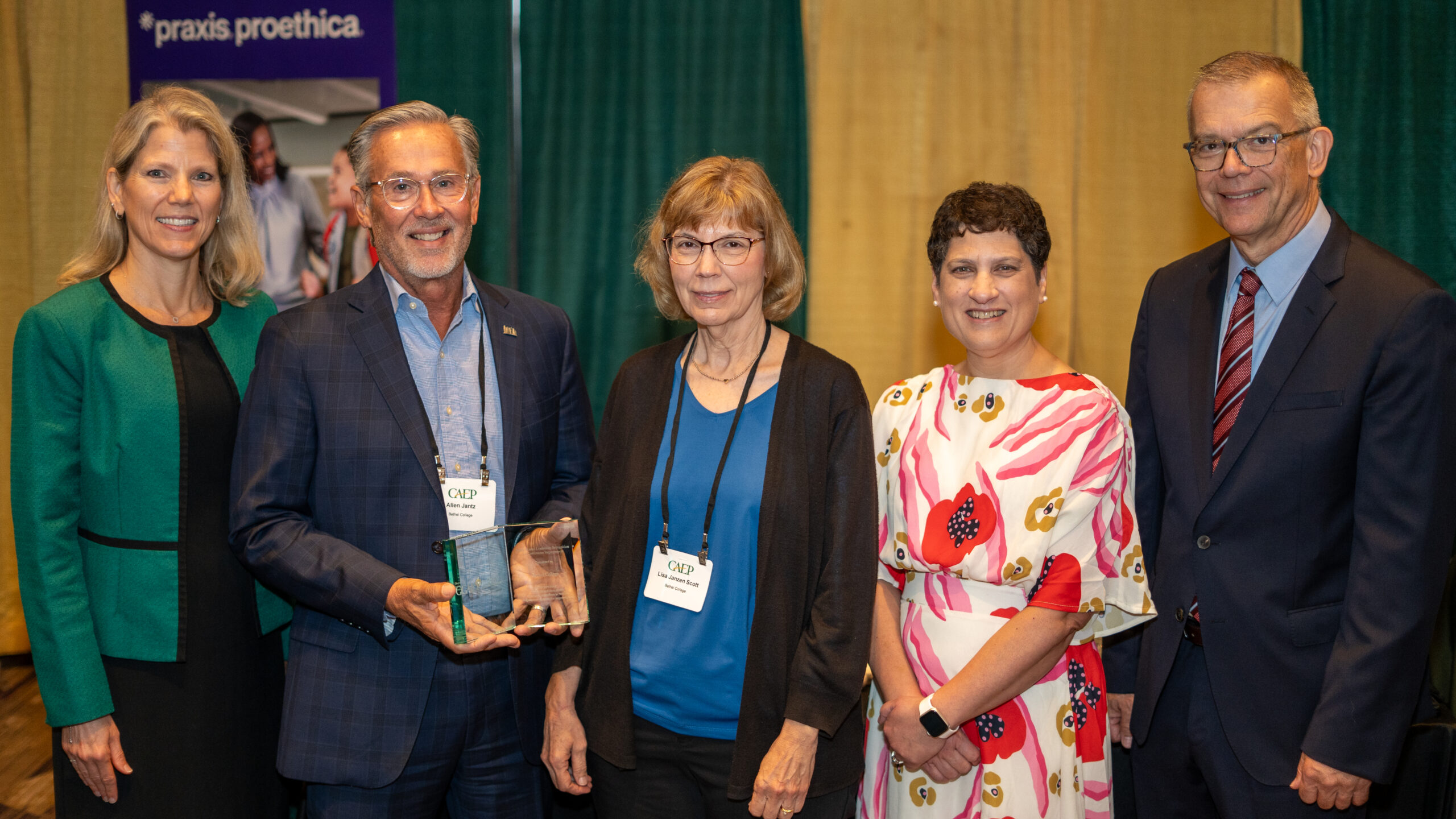 Five people in a line at an awards ceremony; second from left is holding the award.